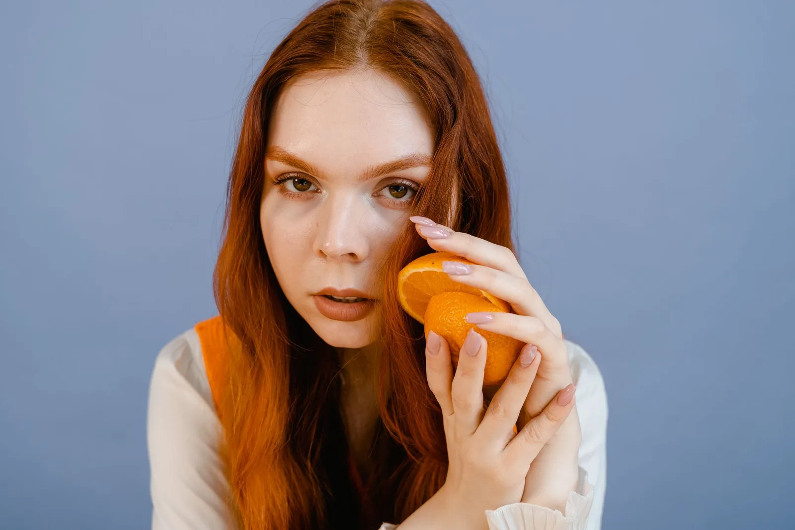 A thoughtful woman with long red hair holds a sliced orange against a blue background.