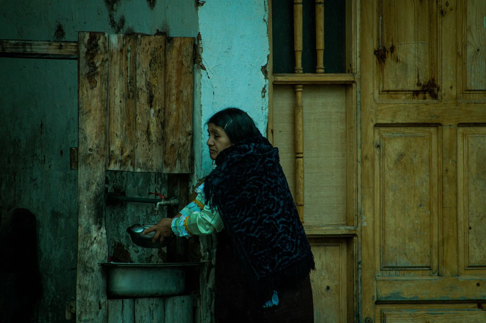 A woman washes her hands at an outdoor rustic sink against a weathered wooden wall.