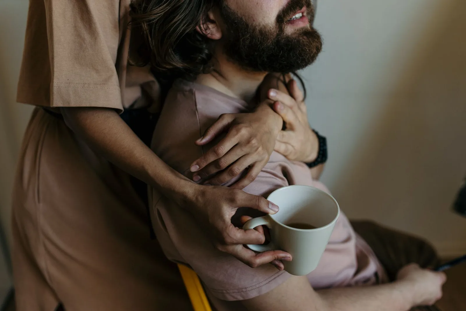 Crop unrecognizable young woman with cup of coffee in casual dress hugging bearded boyfriend from behind while spending time together at hom