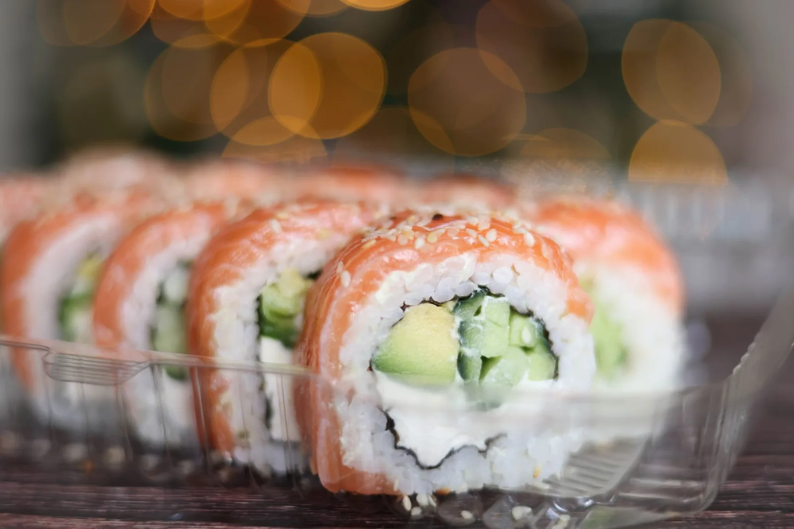 Delicious sushi roll topped with fresh salmon, filled with avocado and cream cheese, in clear plastic tray.