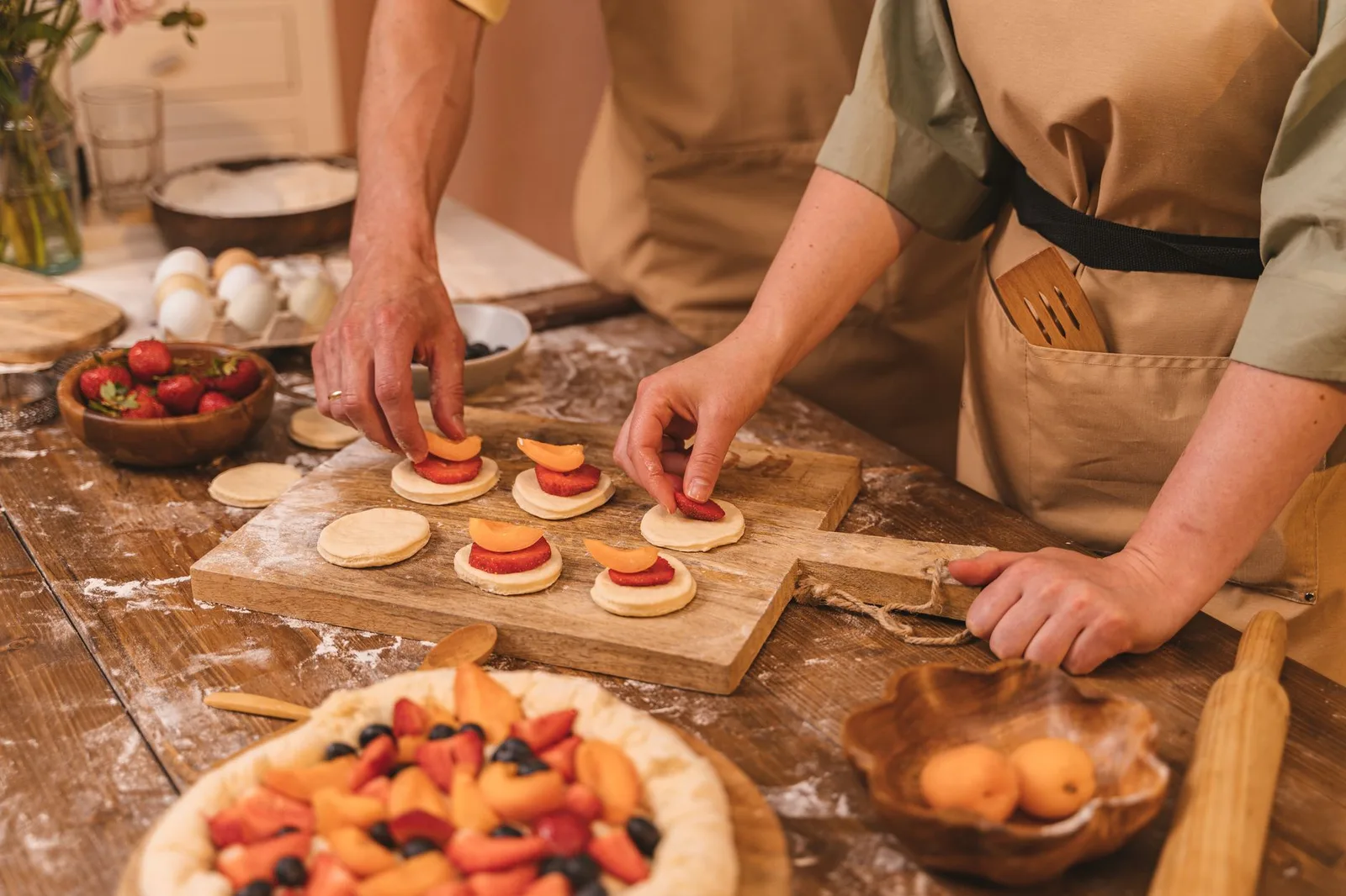 Two chefs arrange sliced fruit on pastry dough for baking. Hands-on cooking in a cozy kitchen.