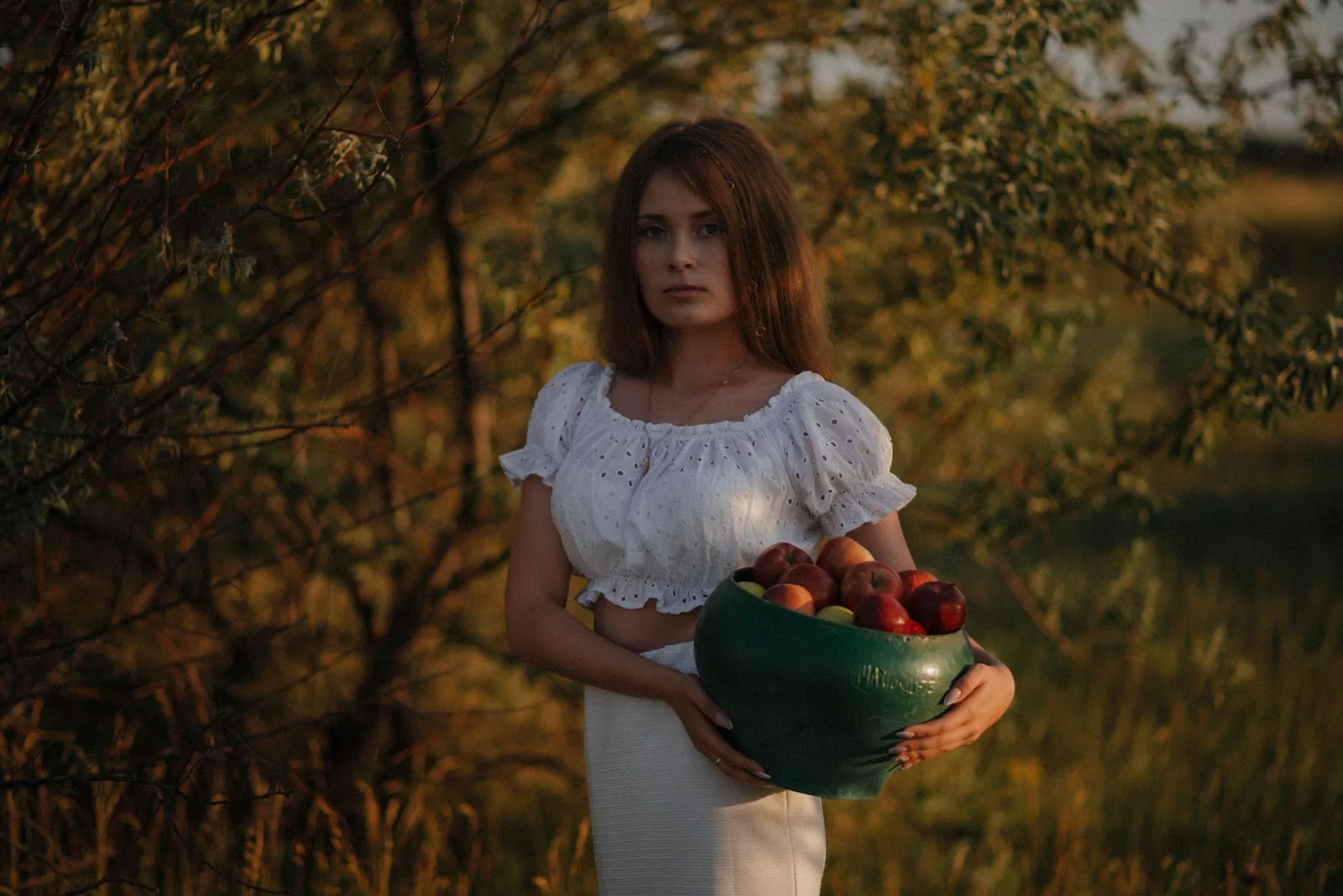 Water drinken en afvallen: hoeveel heb je per dag nodig? — Young woman in a white top holding a bowl of apples in an orchard at sunset.